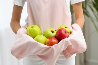 Photo of Woman holding pile of apples with hem of apron indoors, closeup