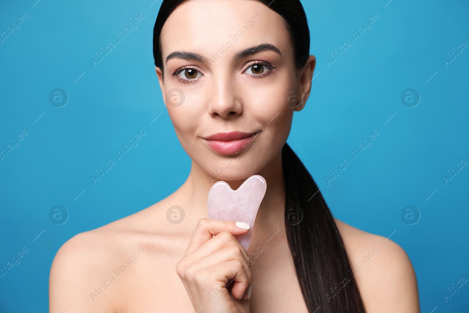 Photo of Beautiful young woman doing facial massage with gua sha tool on blue background, closeup