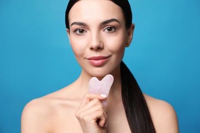 Photo of Beautiful young woman doing facial massage with gua sha tool on blue background, closeup