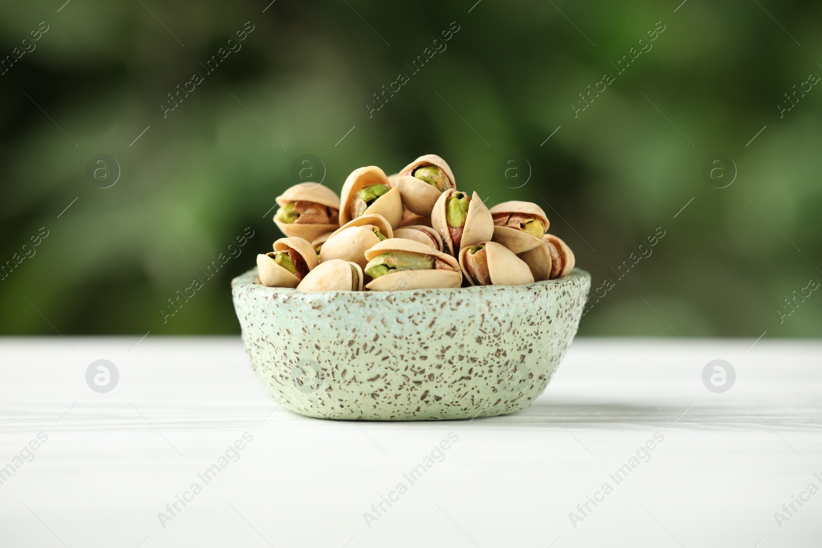 Photo of Tasty pistachios in bowl on white table against blurred background