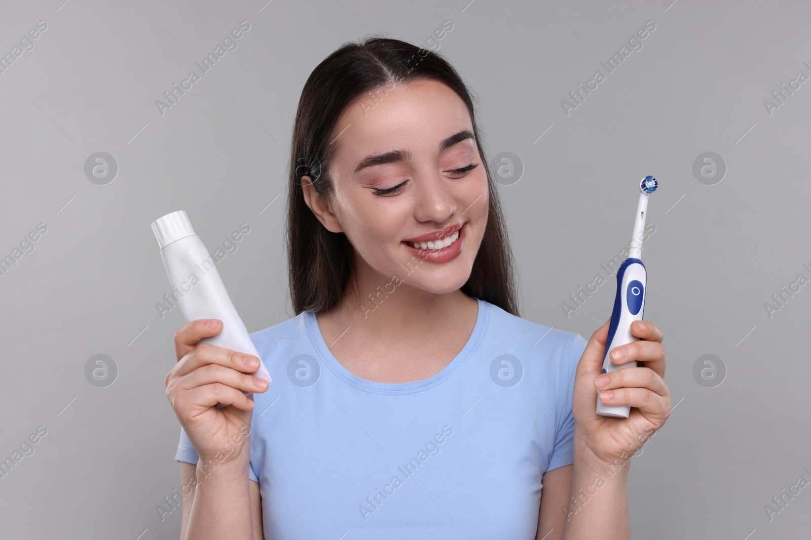 Photo of Happy young woman holding electric toothbrush and tube of toothpaste on light grey background