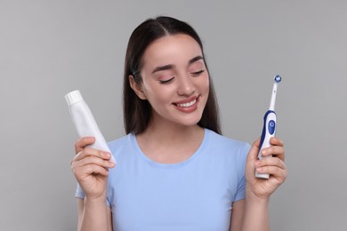 Happy young woman holding electric toothbrush and tube of toothpaste on light grey background