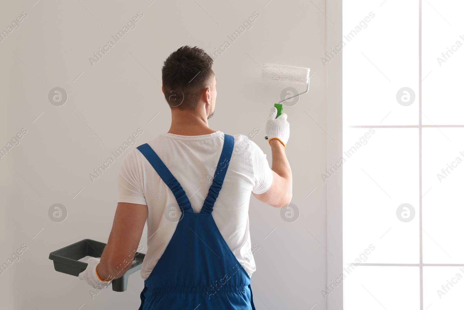 Photo of Man painting wall with white dye indoors, back view