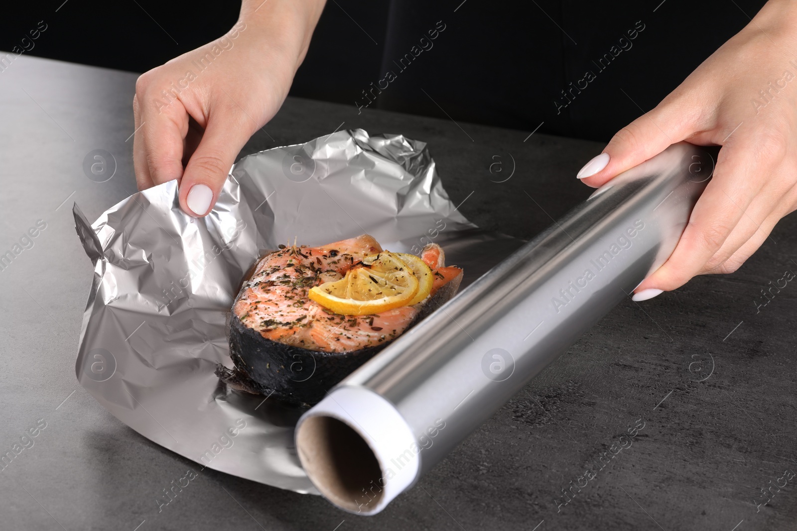 Photo of Woman wrapping tasty salmon with lemon and spices in aluminum foil at grey textured table, closeup