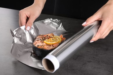 Woman wrapping tasty salmon with lemon and spices in aluminum foil at grey textured table, closeup