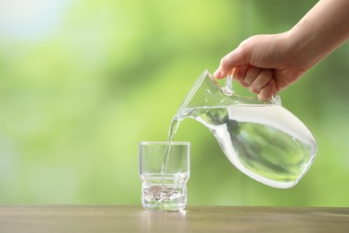 Woman pouring fresh water from jug into glass at wooden table against blurred green background, closeup. Space for text