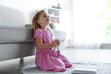 Cute little girl with beads praying in living room. Space for text