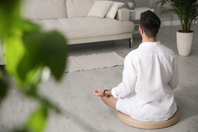 Young man meditating on straw cushion at home, back view