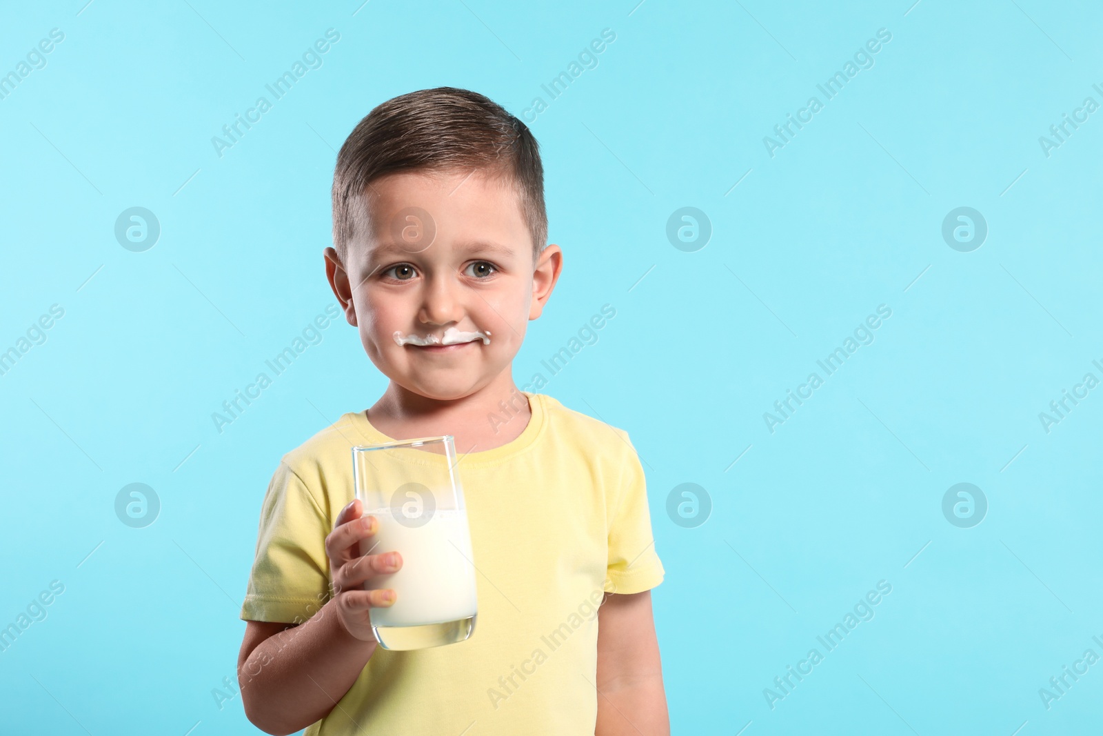 Photo of Cute boy with milk mustache holding glass of tasty dairy drink on light blue background, space for text