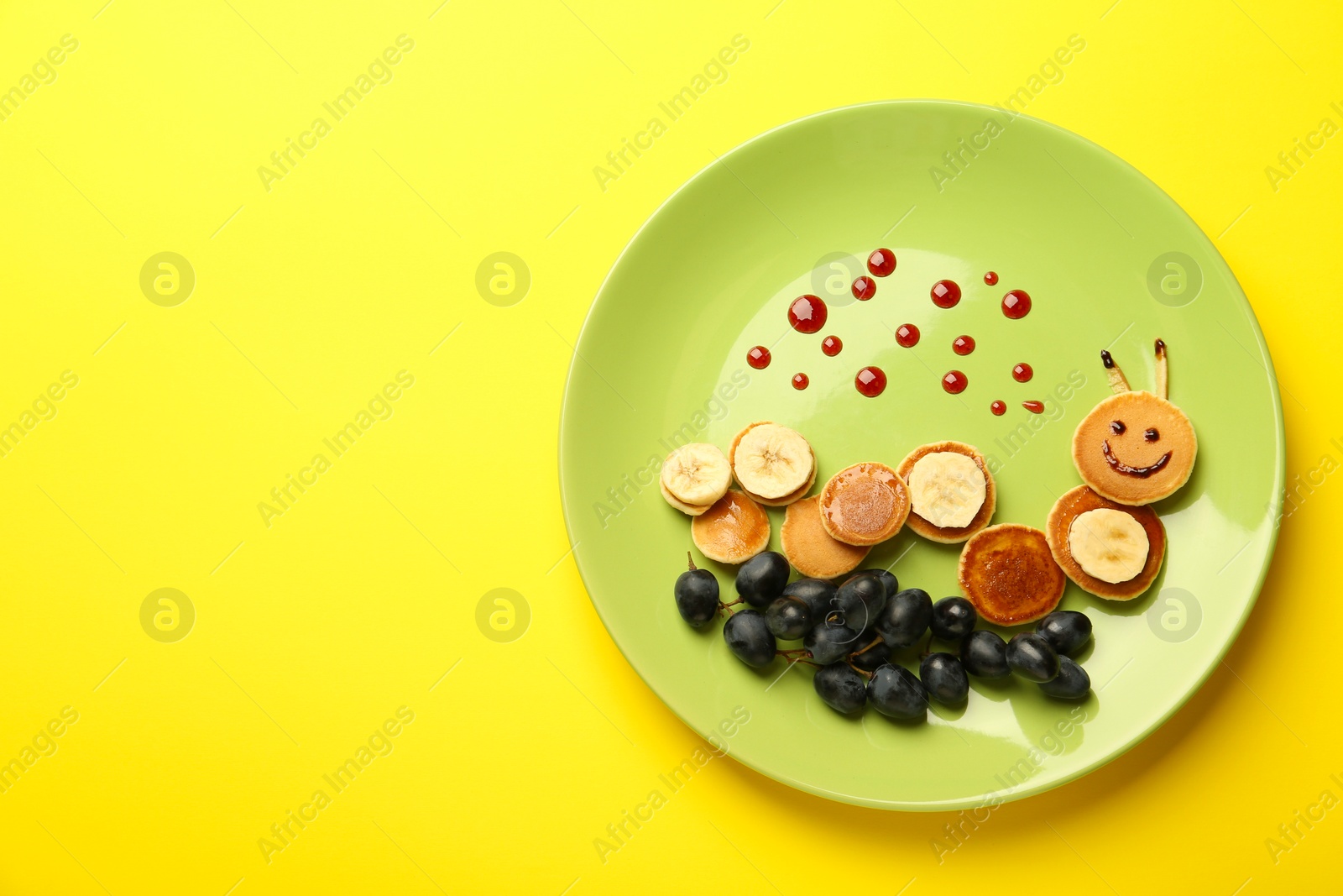 Photo of Creative serving for kids. Plate with cute caterpillar made of pancakes, grapes and banana on yellow background, top view. Space for text