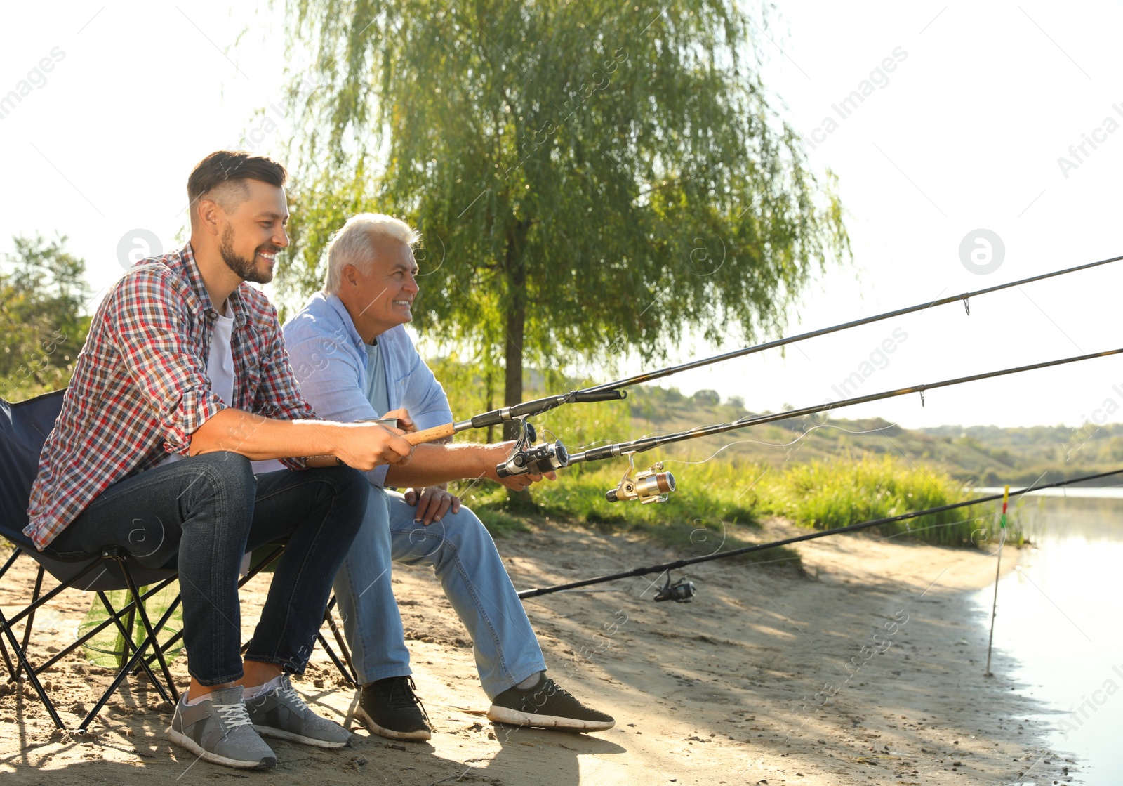 Photo of Father and adult son fishing together from riverside on sunny day