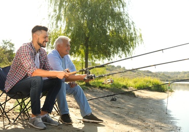 Photo of Father and adult son fishing together from riverside on sunny day