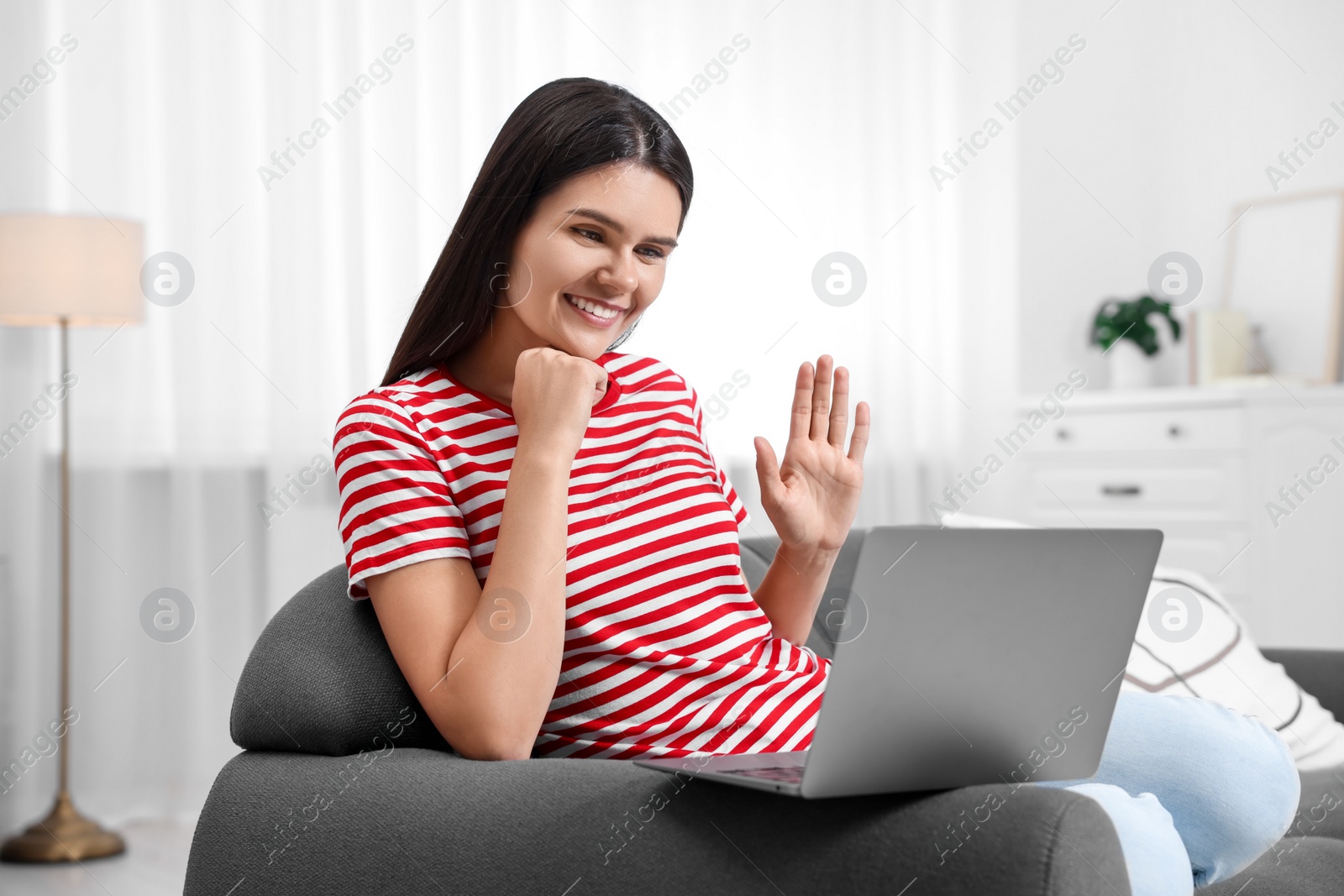 Photo of Happy young woman having video chat via laptop and waving hello on sofa in living room