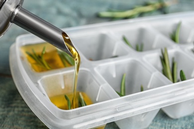 Photo of Pouring olive oil into ice cube tray with rosemary on table, closeup