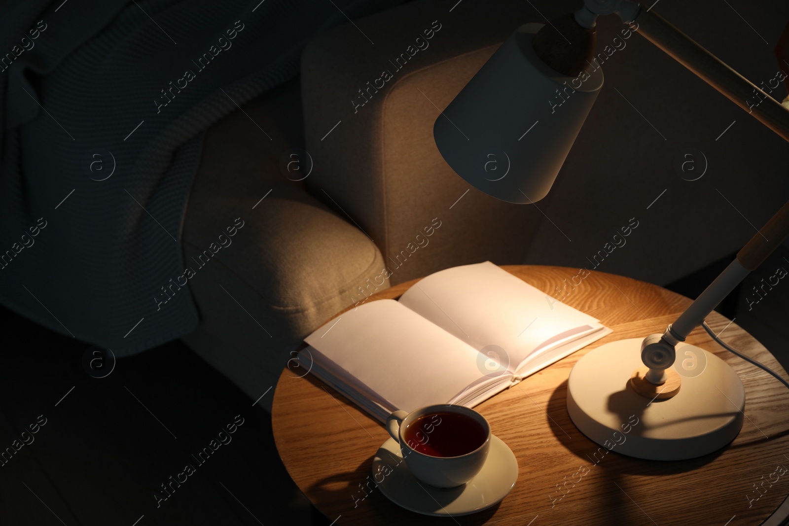Photo of Stylish lamp, book and cup of tea on side table near sofa indoors