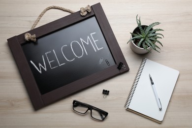 Photo of Blackboard with word Welcome, stationery, plant and glasses on wooden table, flat lay