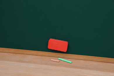 Photo of Colorful chalks on wooden table near green board with duster. Space for text