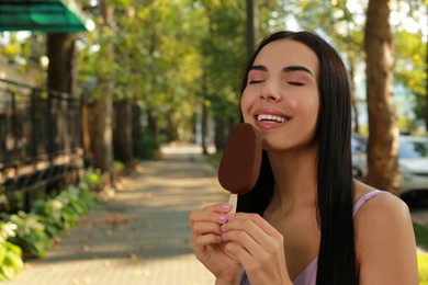 Photo of Beautiful young woman eating ice cream glazed in chocolate on city street, space for text