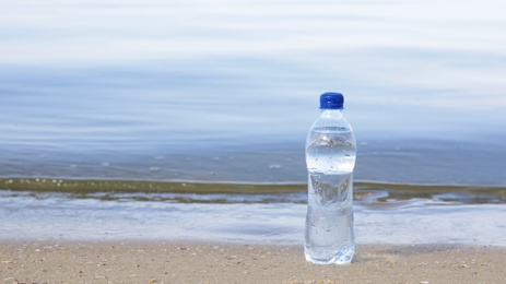 Plastic bottle with refreshing drink on sandy beach near sea, space for text. Hot summer day