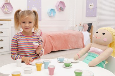 Photo of Cute little girl playing tea party with doll at table in room, space for text