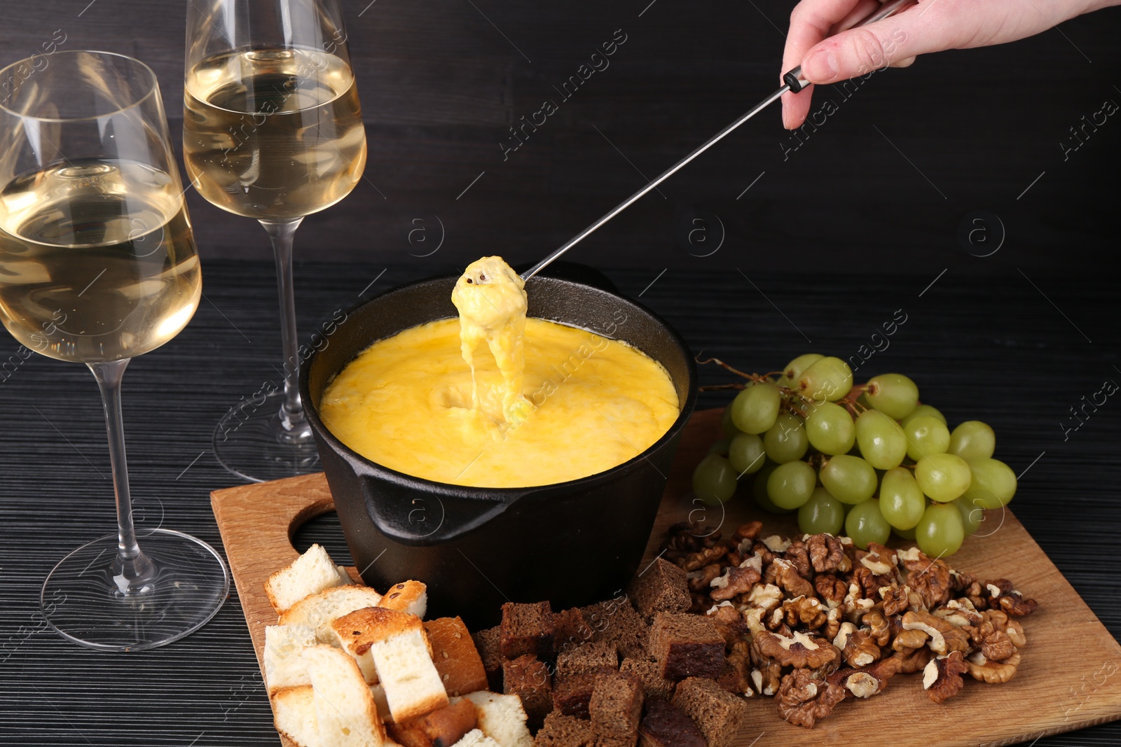 Photo of Woman dipping piece of bread into fondue pot with melted cheese at black wooden table, closeup