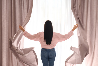 Woman opening window curtains at home in morning, back view