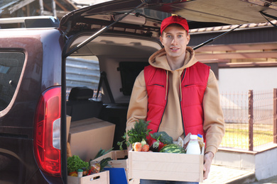 Photo of Courier holding crate with products near car outdoors. Food delivery service