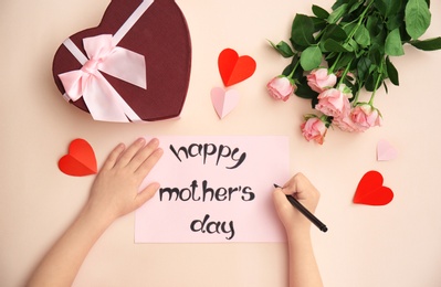 Photo of Child writing phrase "HAPPY MOTHER'S DAY" on card. Gift and roses on table