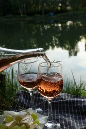 Pouring delicious rose wine into glass on picnic blanket near lake, closeup