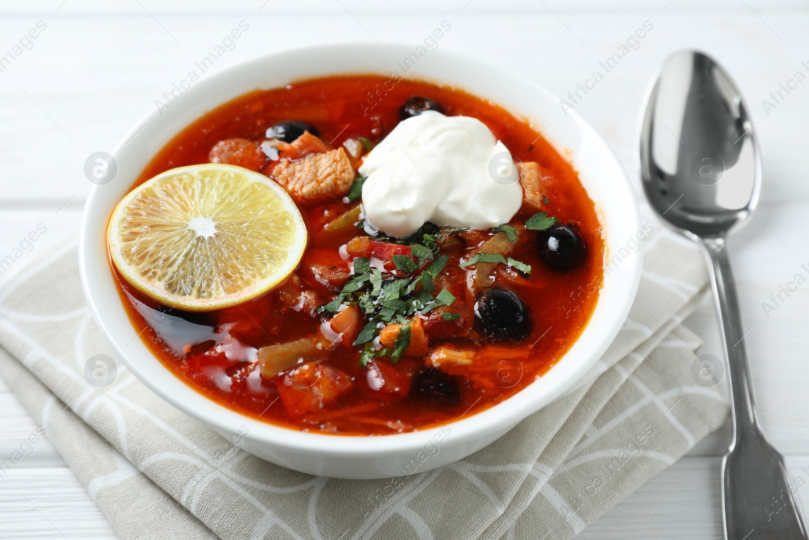 Photo of Meat solyanka soup with sausages, olives, vegetables and spoon on white wooden table, closeup