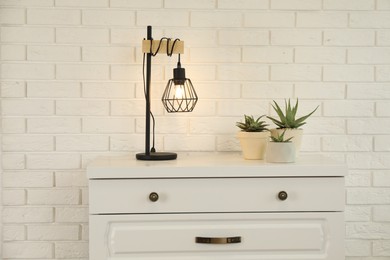 Photo of Modern chest of drawers with houseplants and lamp near white brick wall