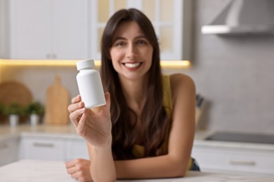 Beautiful woman with bottle of vitamin pills at white table in kitchen