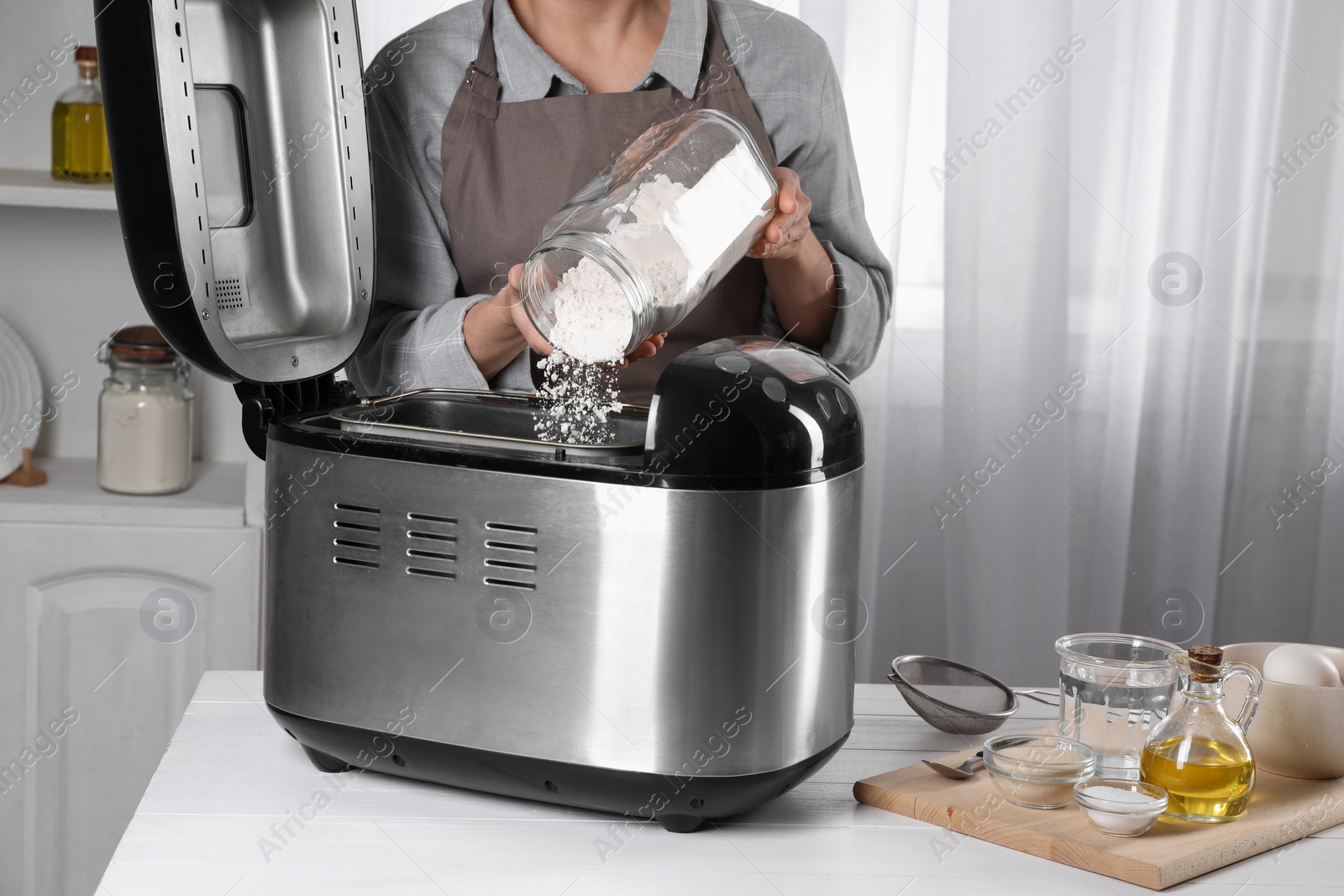 Photo of Woman adding flour into breadmaker at white wooden table indoors, closeup