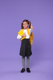 Photo of Smiling schoolgirl with backpack and book showing thumbs up on violet background