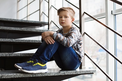 Photo of Depressed little boy sitting on stairs indoors. Time to visit child psychologist