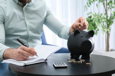 Businessman with notebook, piggy bank and money at table indoors, closeup