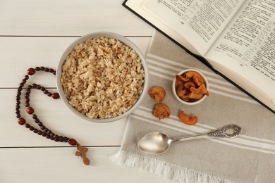 Rosary beads, oatmeal porridge, dried apples and Bible on white wooden table, flat lay. Lent season