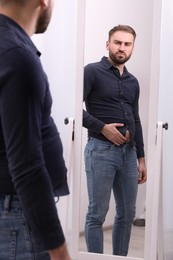 Upset man wearing tight shirt in front of mirror at home. Overweight problem