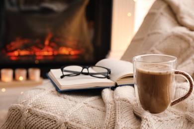 Cup of coffee, book and glasses near fireplace indoors, space for text. Cozy atmosphere