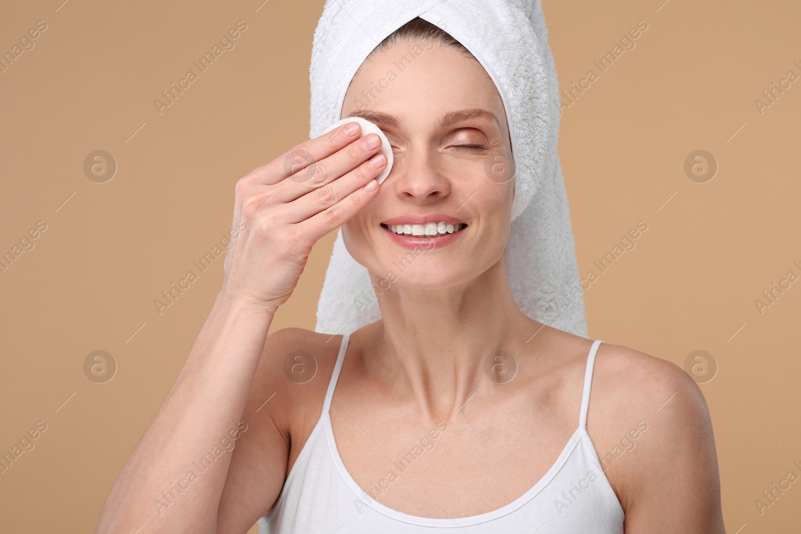 Photo of Beautiful woman in terry towel removing makeup with cotton pad on beige background