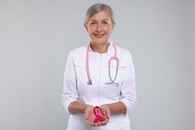 Photo of Doctor with stethoscope holding pink ribbon on light grey background. Breast cancer awareness