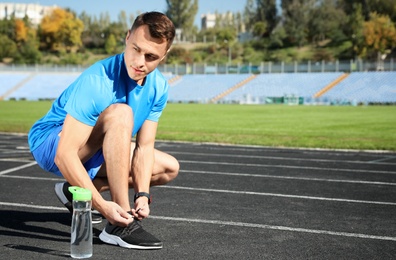 Photo of Sporty man tying shoelaces near bottle of water at stadium on sunny day. Space for text