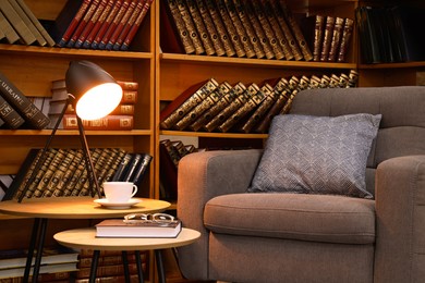 Book with glasses, table lamp and cup of drink near comfortable armchair in cozy home library