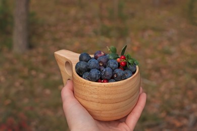 Photo of Woman holding wooden mug full of fresh ripe blueberries and lingonberries in forest, closeup