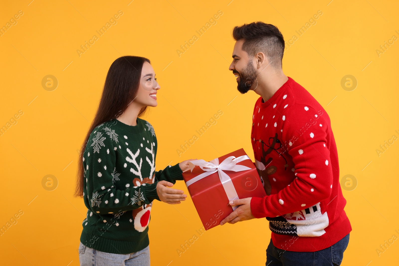 Photo of Couple in Christmas sweaters. Young man presenting gift to his woman on orange background