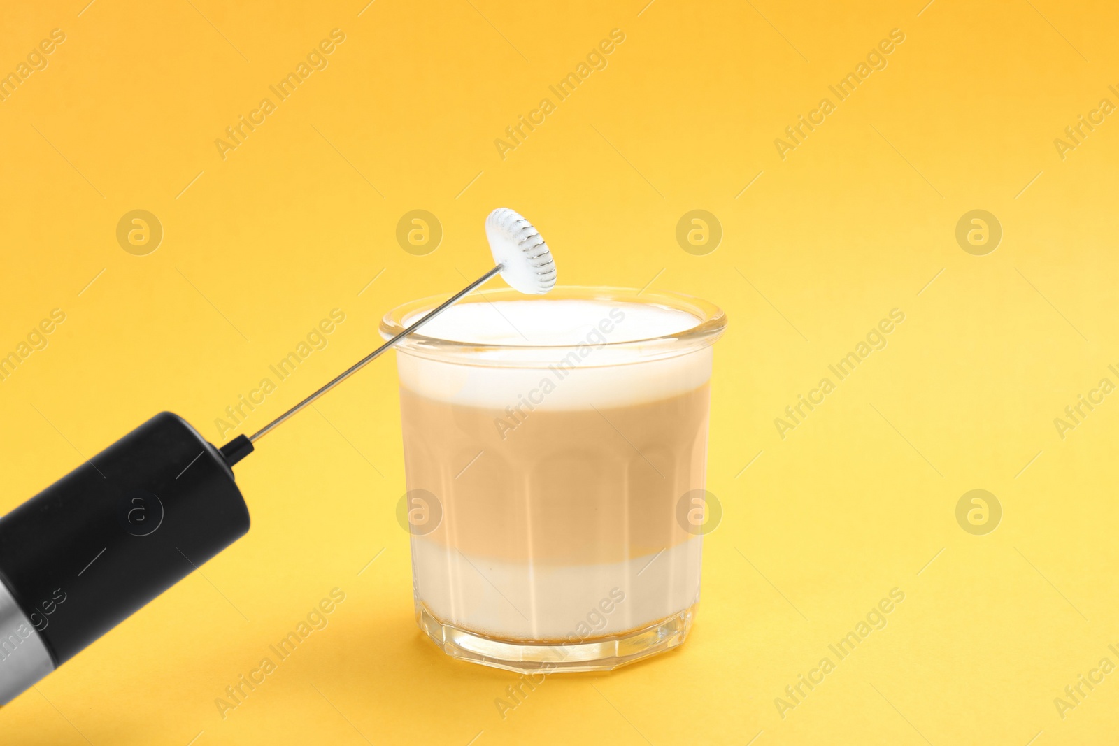 Photo of Mini mixer (milk frother) and tasty cappuccino in glass on yellow background