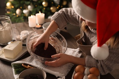 Photo of Little child in Santa hat making Christmas cookies at table, closeup