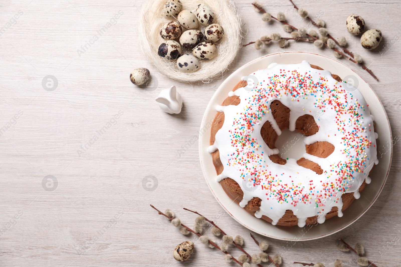 Photo of Glazed Easter cake with sprinkles, decorative bunny, quail eggs and willow branches on white wooden table, flat lay. Space for text