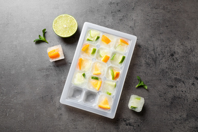 Photo of Ice cube tray with frozen fruits and mint on grey table, flat lay