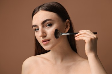 Photo of Beautiful woman with freckles applying makeup with brush on brown background